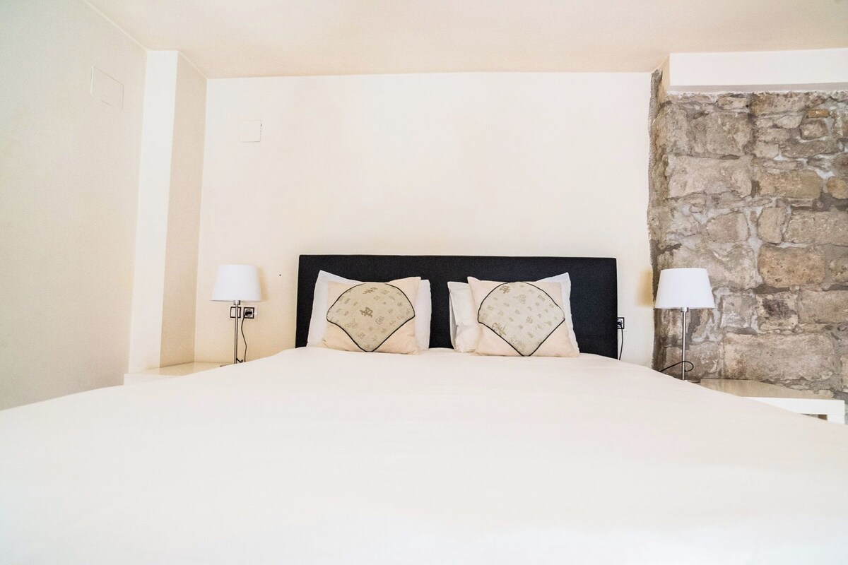 A bed with a black headboard, two bedside lamps, and a stone wall.