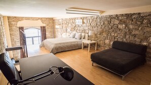 A bedroom with a stone wall, a bed, a bedside table with a lamp, a chair, and a desk with a lamp. - Urbi Apartments (Manresa)