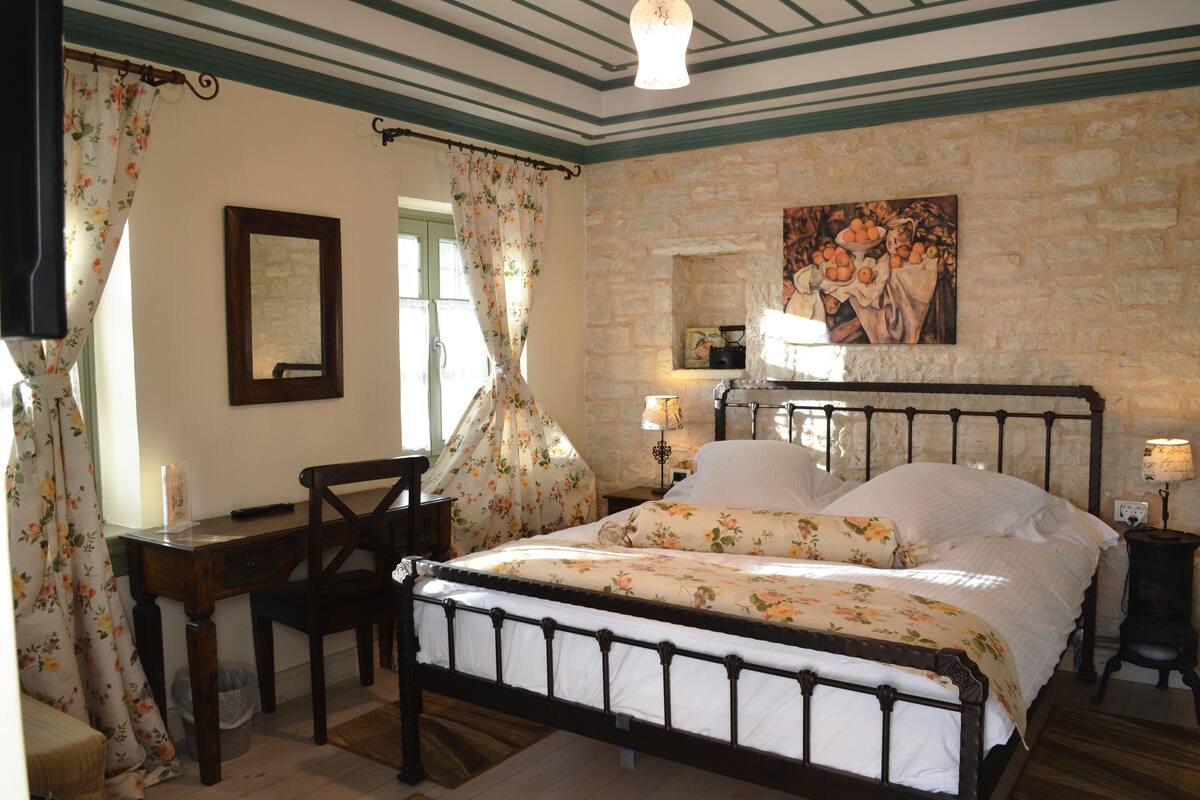 Een slaapkamer met een stenen muur, een bed met bloemenbeddels, een houten bureau en een nachtkastje met een lamp.