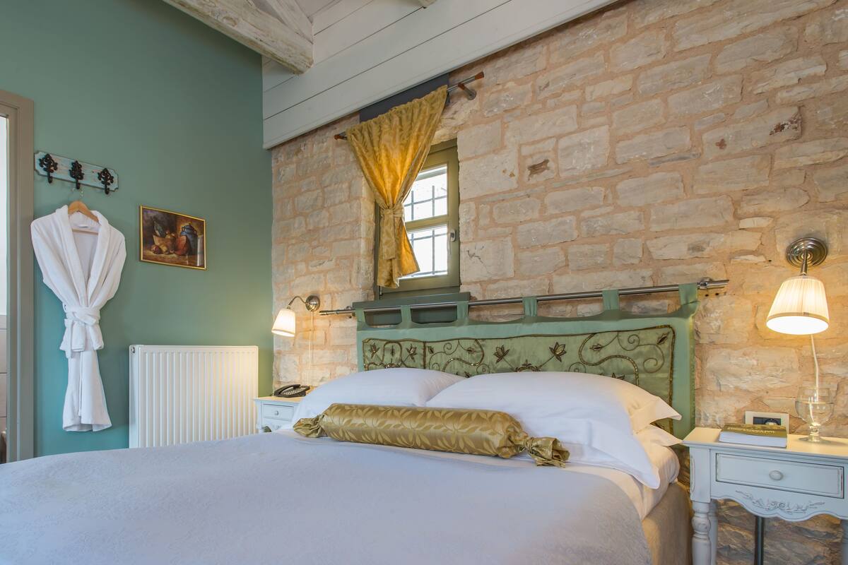 Een slaapkamer met een stenen muur, een bed met een gouden kussen, een nachtkastje met een lamp, en een badkamer met een wastafel en een badjas.