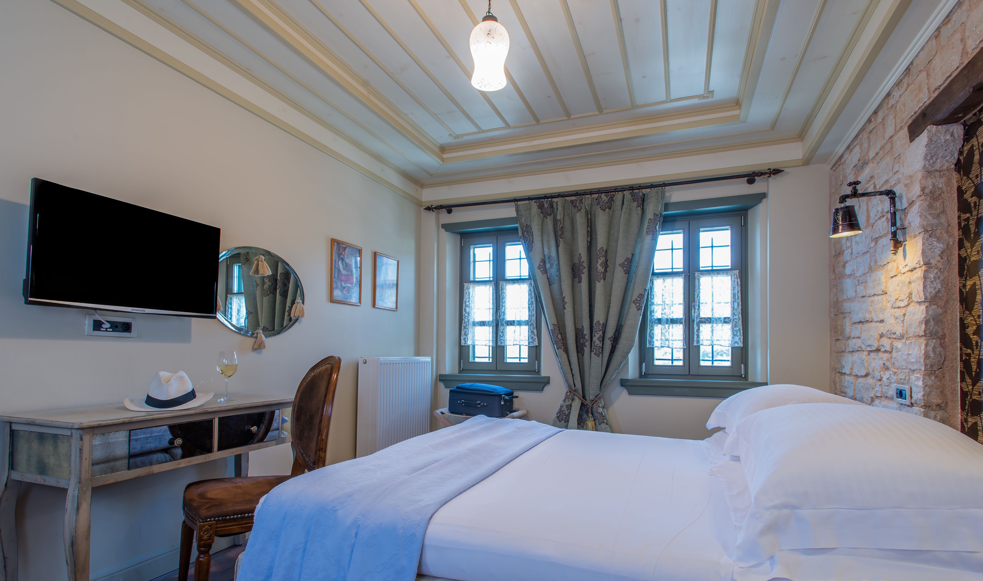 A hotel room with a bed, a desk with a chair, a television, a mirror, and two windows with curtains.
