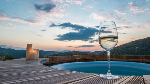 Un verre de vin blanc sur une terrasse en bois donnant sur une piscine, avec des montagnes en arrière-plan.