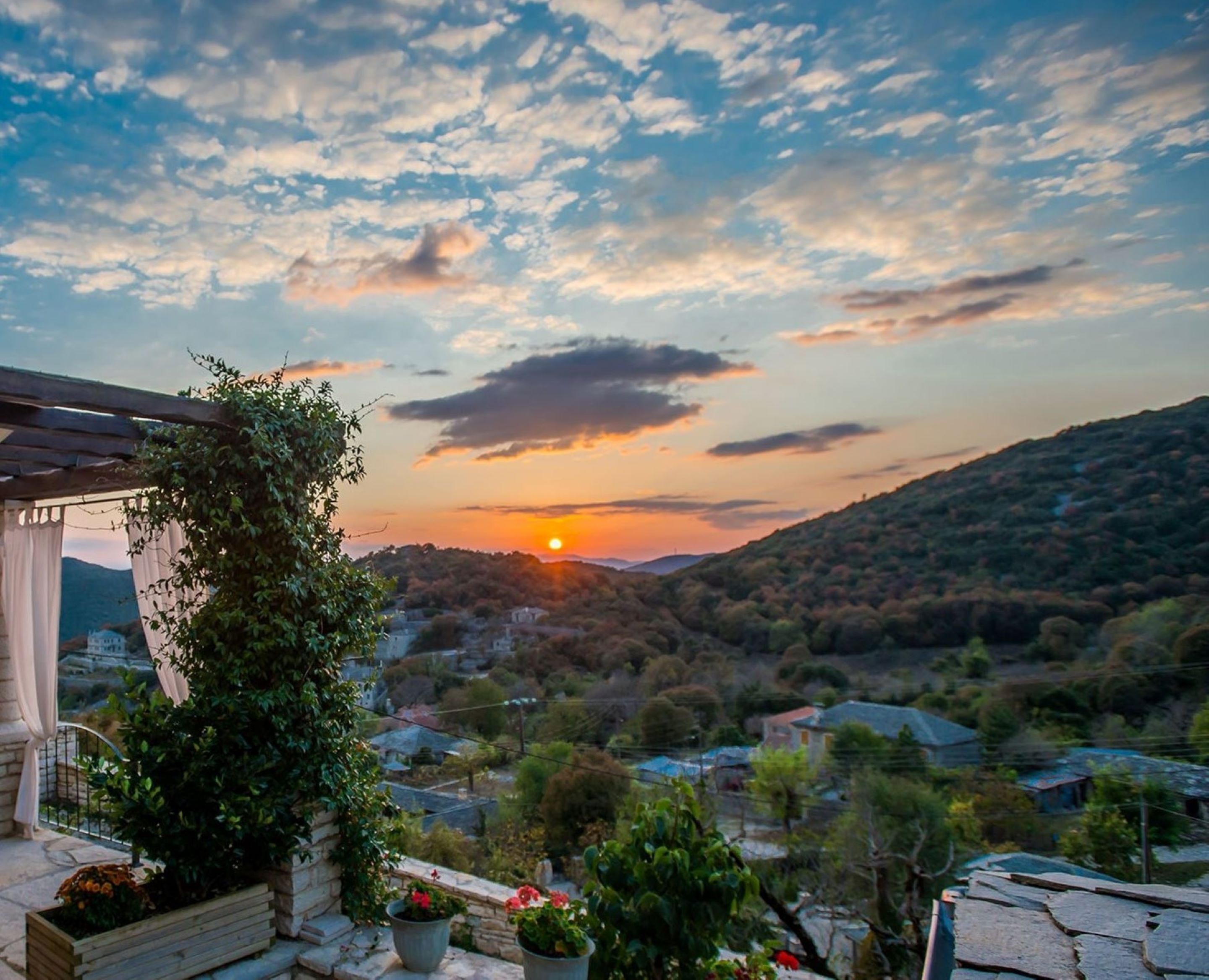 A sunset view from a balcony with a pergola, potted plants, and a stone wall.