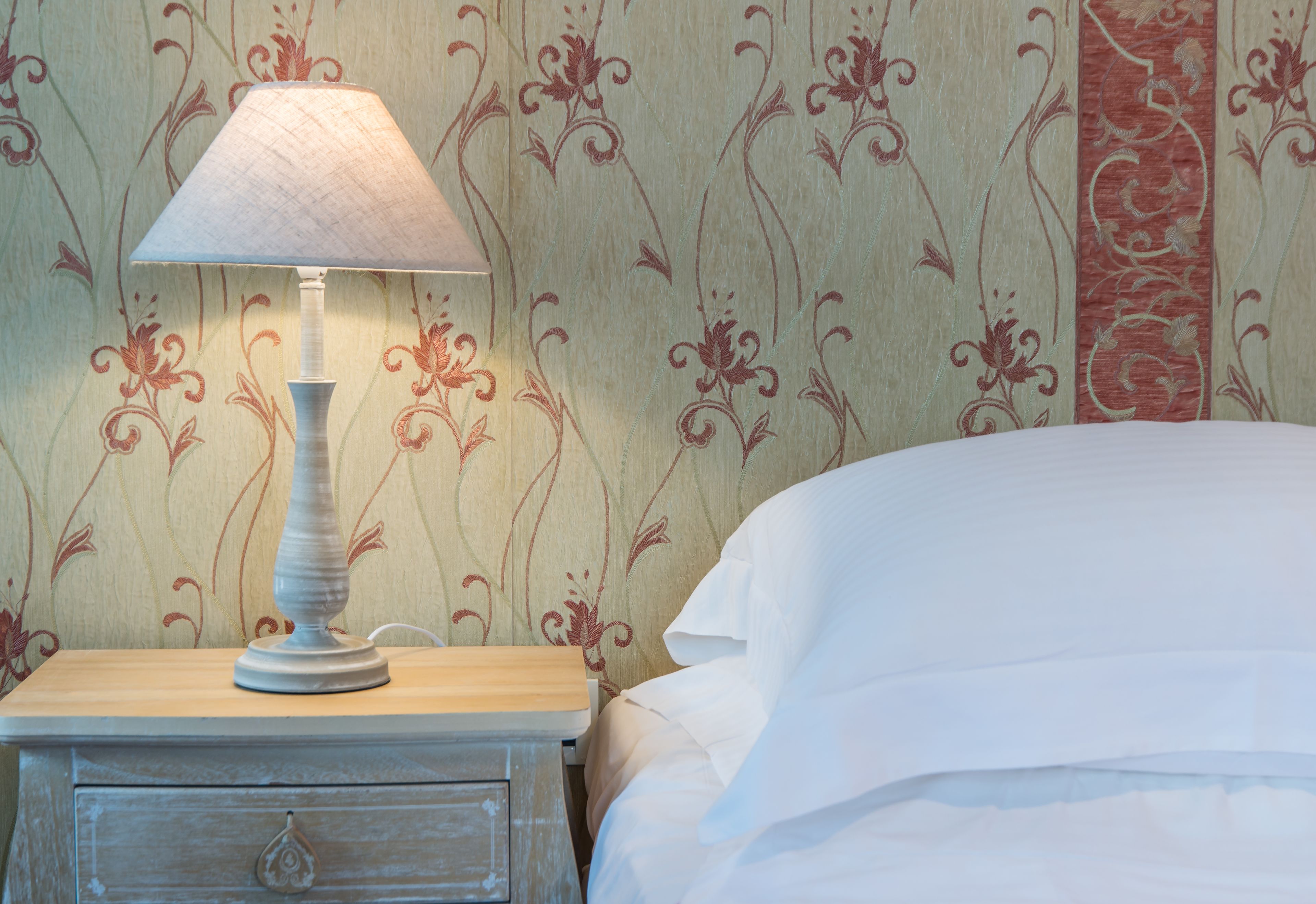 Una cama bien hecha con ropa blanca, una lámpara blanca sobre una mesita de noche de madera, y una pared con papel tapiz floral.