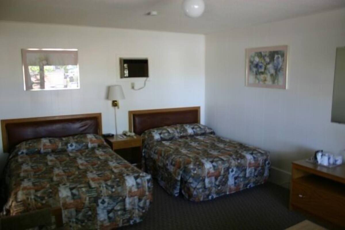 A hotel room with two beds, a bedside table with a lamp, a small desk with a chair, and a painting on the wall.
