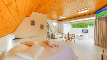 Quarto de hotel com teto de madeira, cama com roupa branca, área de estar com mesa e cadeiras, e uma janela grande com vista para o verde.
