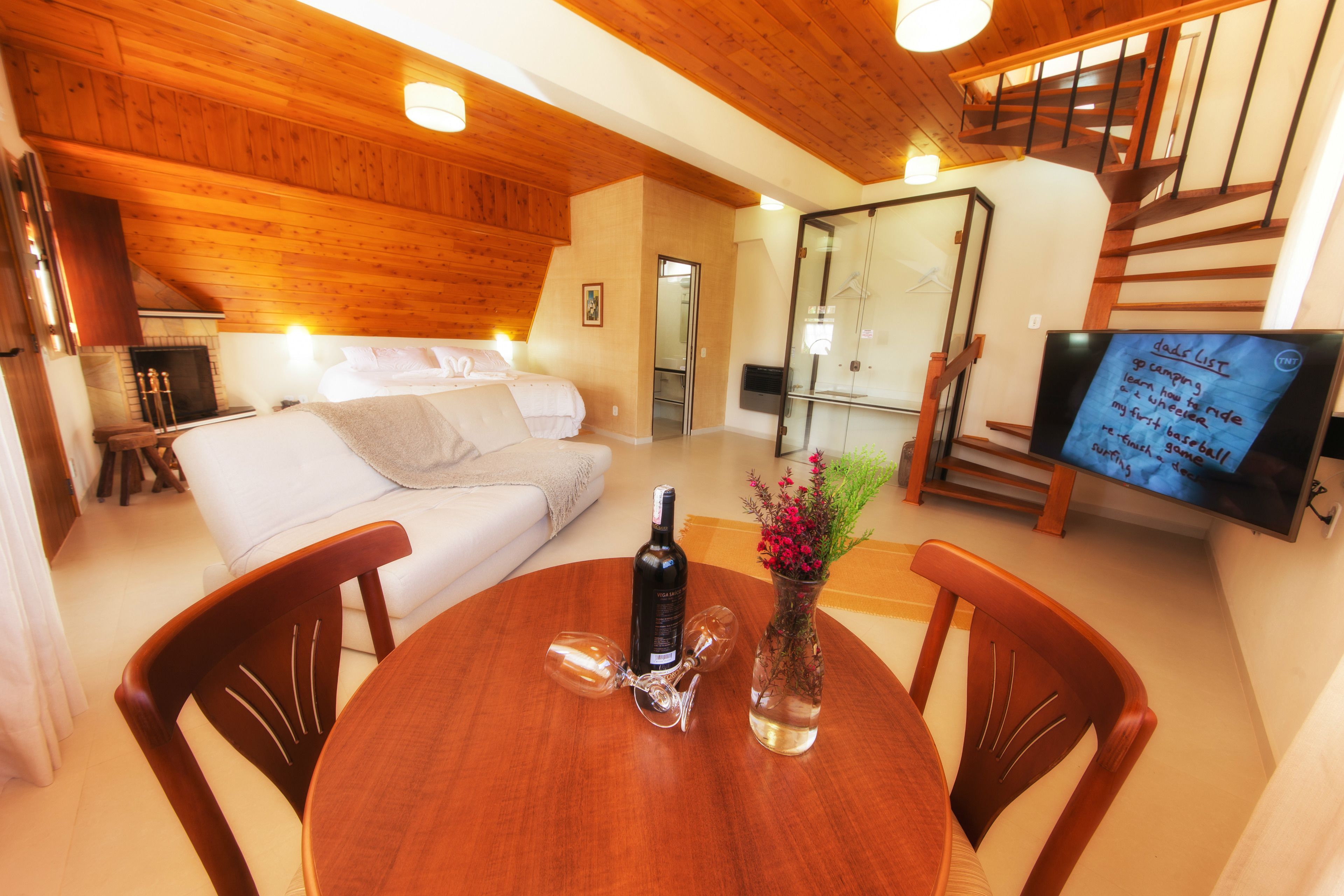 Una sala de estar acogedora con techo de madera, un sofá, una mesa de comedor con sillas, una chimenea y un televisor mostrando una lista.