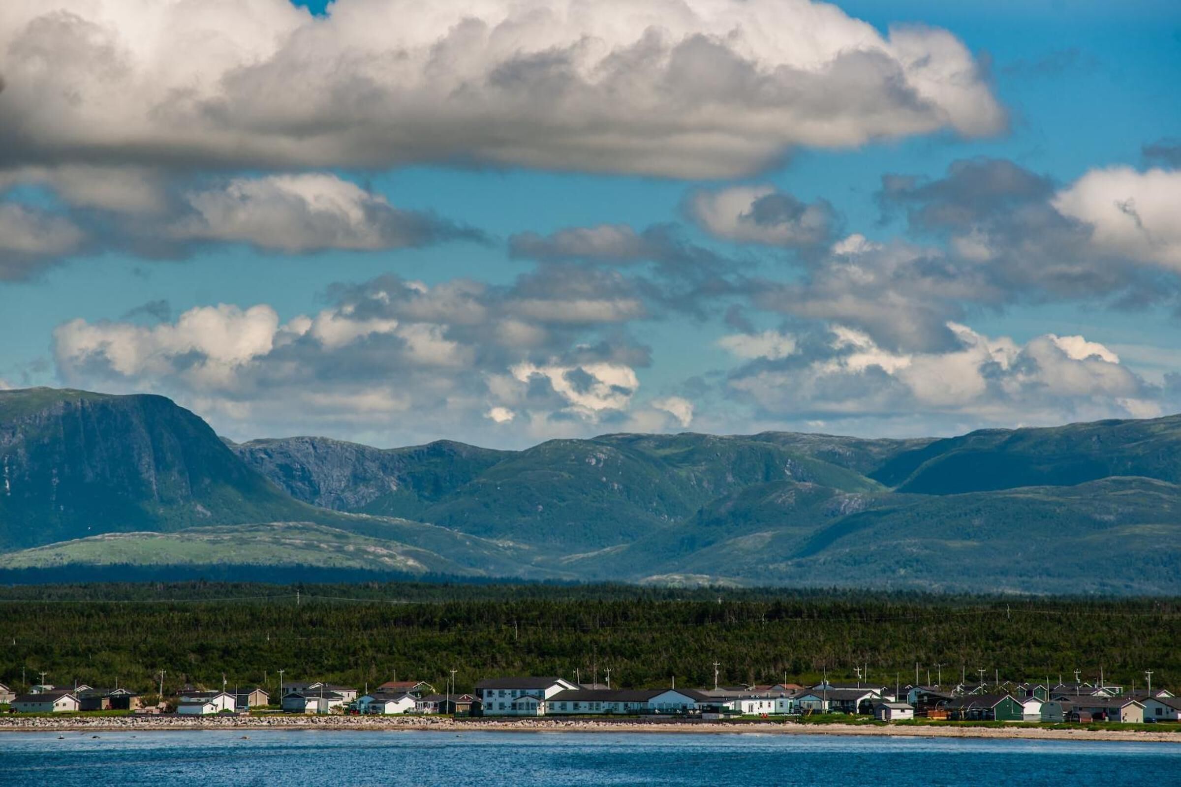A coastal town with houses along the shoreline, a mountain range in the background, and a clear sky with clouds.
