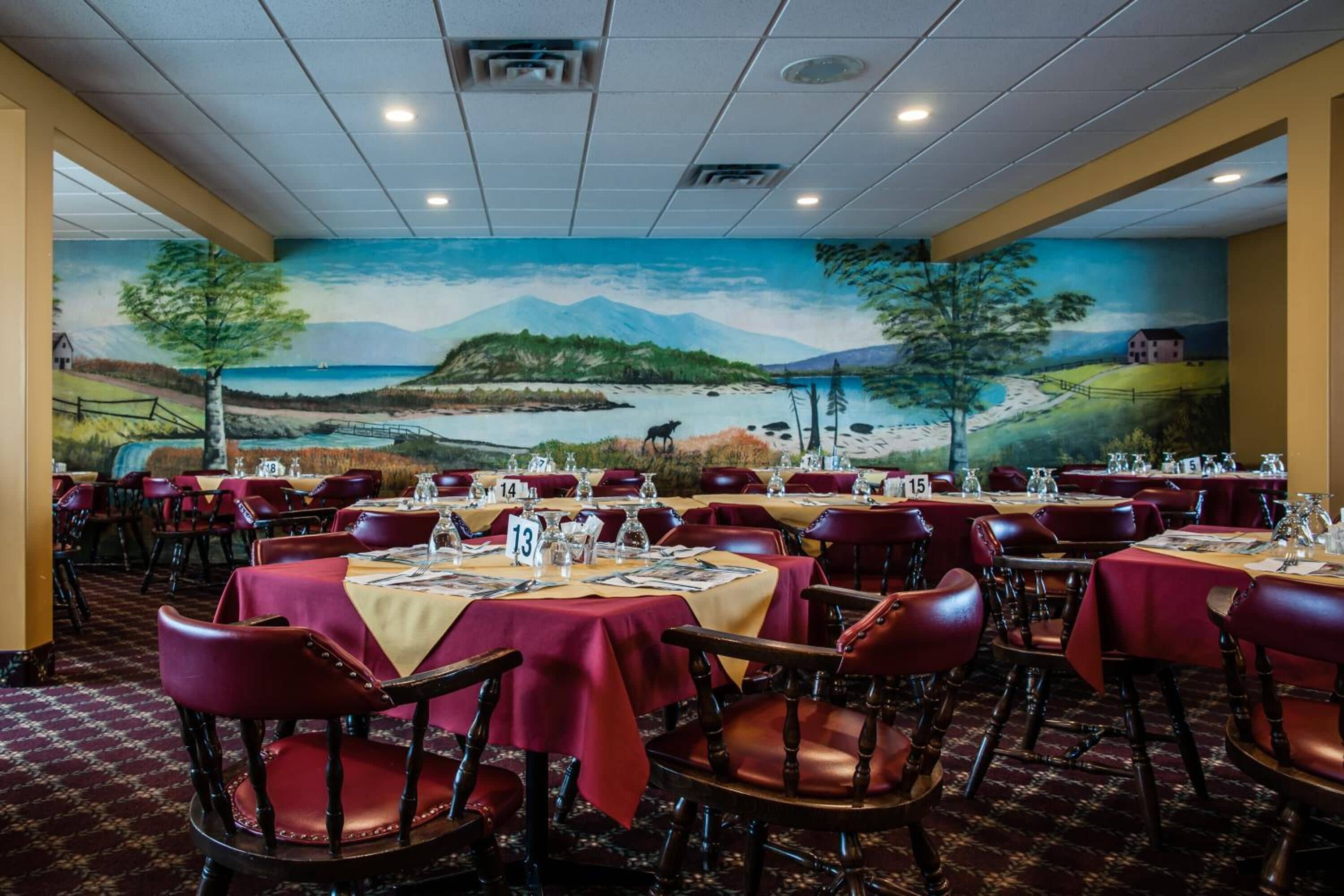 Un espace repas avec des tables dressées pour les convives, des places numérotées et une fresque représentant un paysage sur le mur.