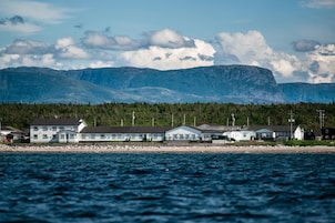 A coastal building complex with multiple structures, a rocky shoreline, and a backdrop of mountains.
