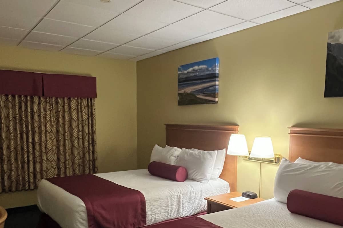 A hotel room with two beds, a headboard, a nightstand, a lamp, a painting, and a window with curtains.
