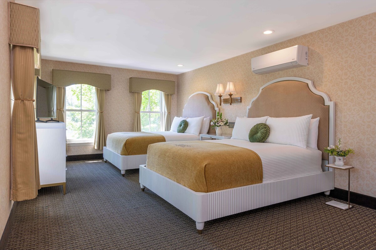 A hotel room with two beds, a large window, a mirror, a bedside table, and a wall-mounted air conditioner.