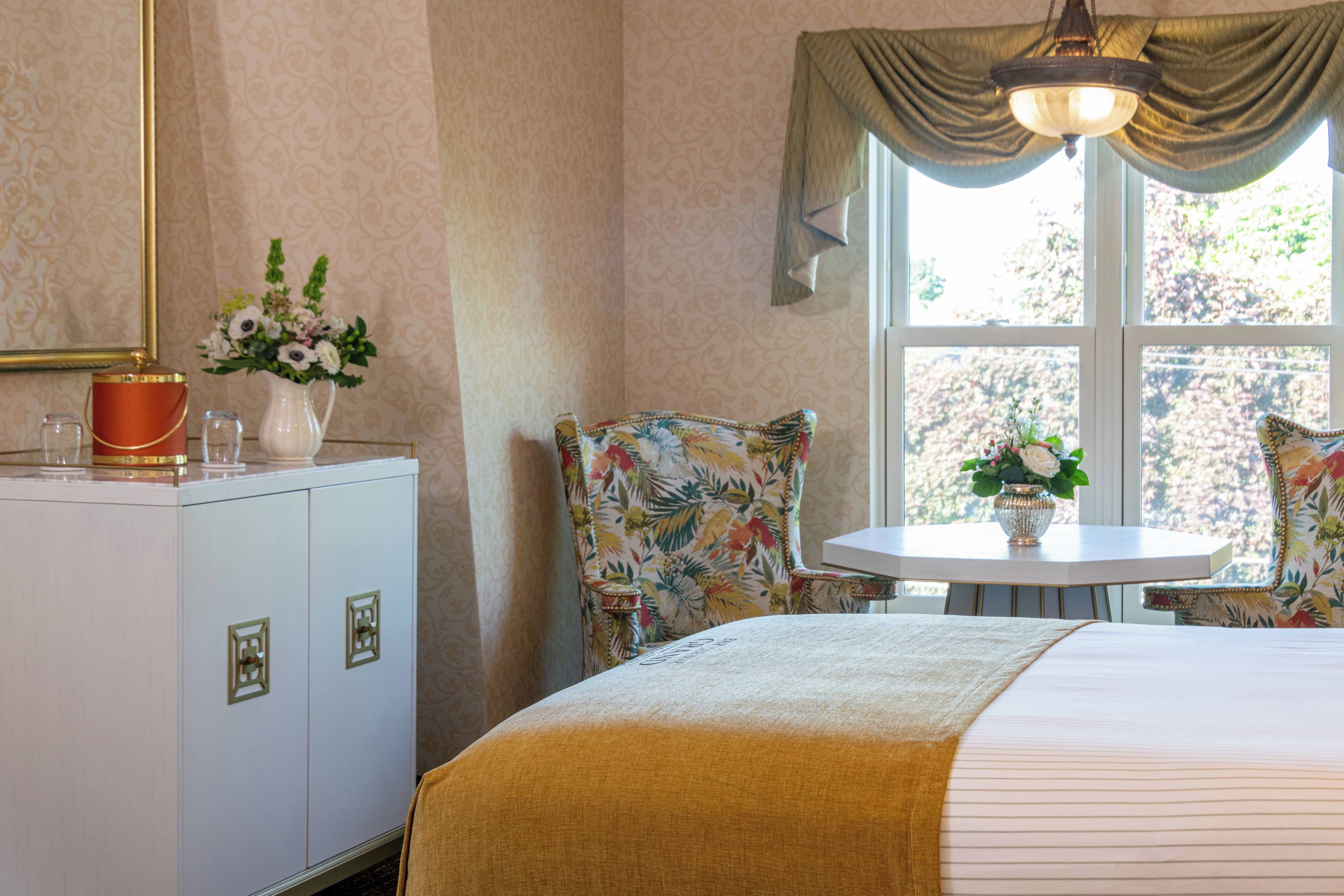 A hotel room with a bed, a small round table with a vase of flowers, two chairs with floral upholstery, a cabinet with a red container, and a window with curtains.