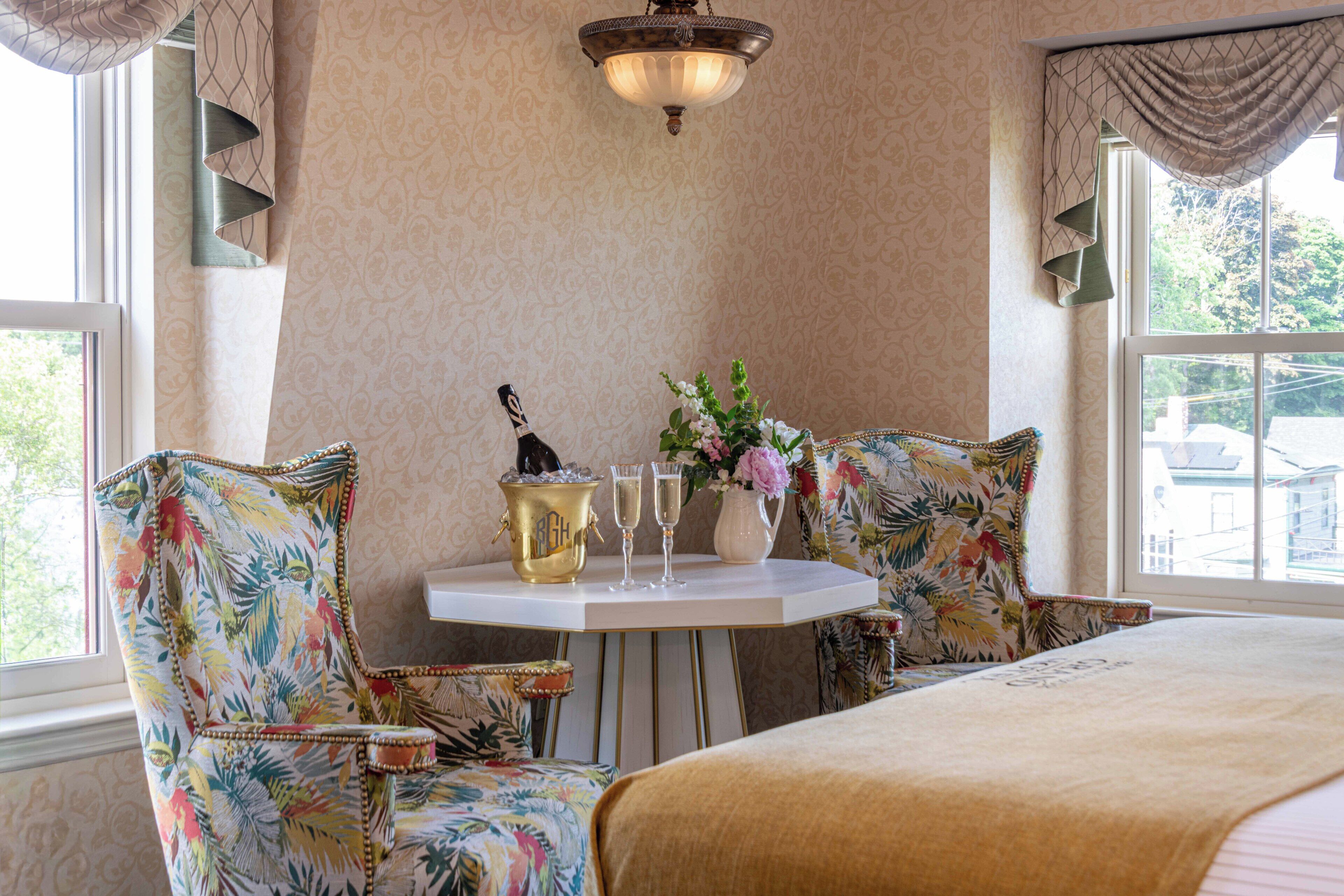 A room with a bed, two floral-patterned armchairs, a small table with a bottle of champagne, two glasses, and a vase of flowers.