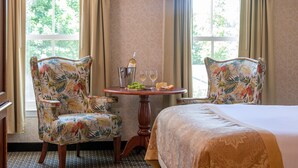 A hotel room with a bed, two floral armchairs, a small table with a bottle, glasses, and a fruit bowl. - Bar Harbor Grand Hotel (Bar Harbor)
