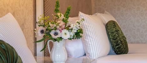 En snyggt bäddad säng med vita sängkläder och en dekorativ kudde, en vit vas med blommor och en grön kudde med struktur.