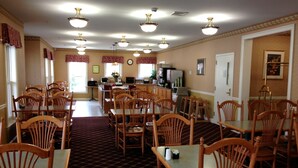 A dining area with multiple wooden tables and chairs, a kitchen area in the background, and a clock on the wall. - Bar Harbor Grand Hotel (Bar Harbor)