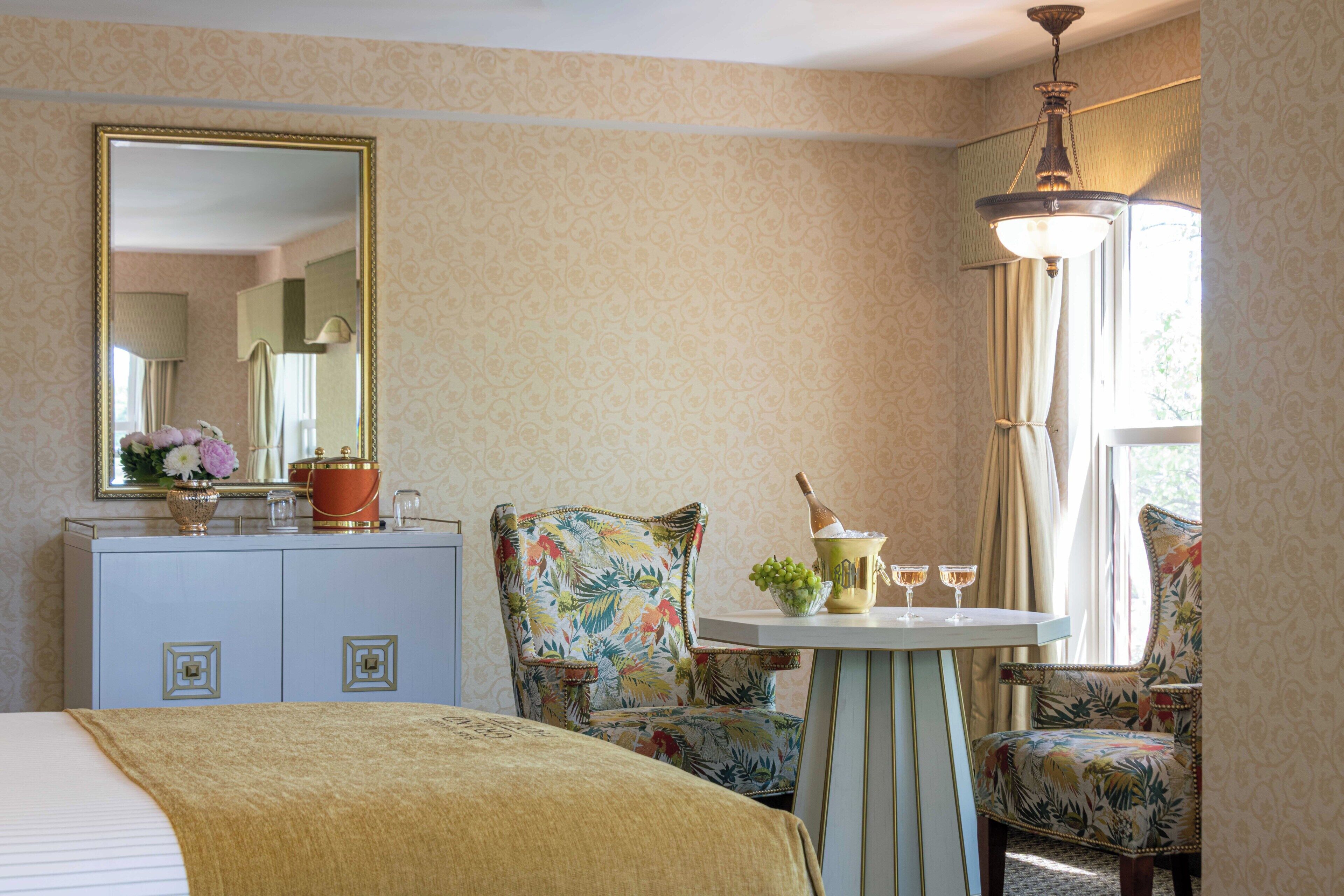 Habitación de hotel con una cama, cómoda, espejo, sillón floral, una mesita con una botella y vasos, y una ventana con cortinas.