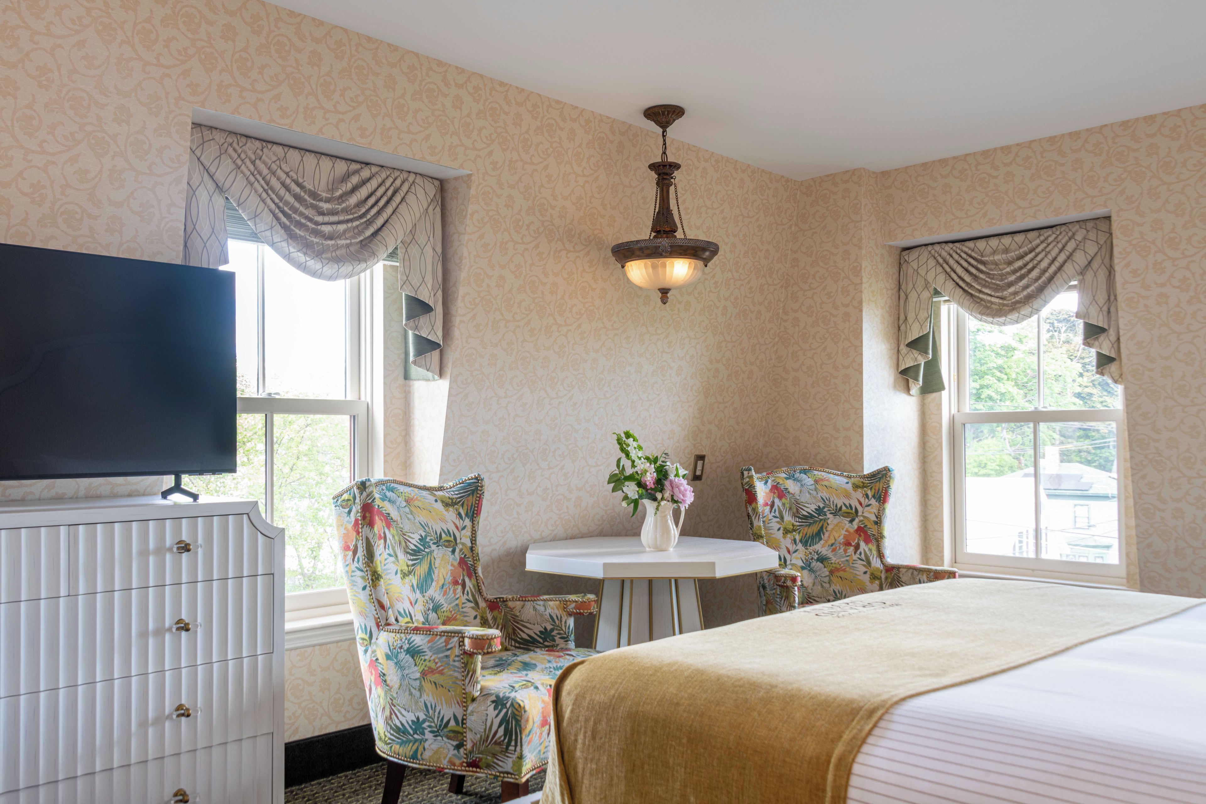 A hotel room with a bed, two chairs, a small table with a vase of flowers, a television, and a window with curtains.