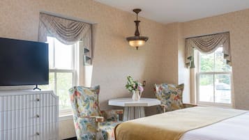 A hotel room with a bed, two chairs, a small table with a vase of flowers, a television, and a window with curtains.