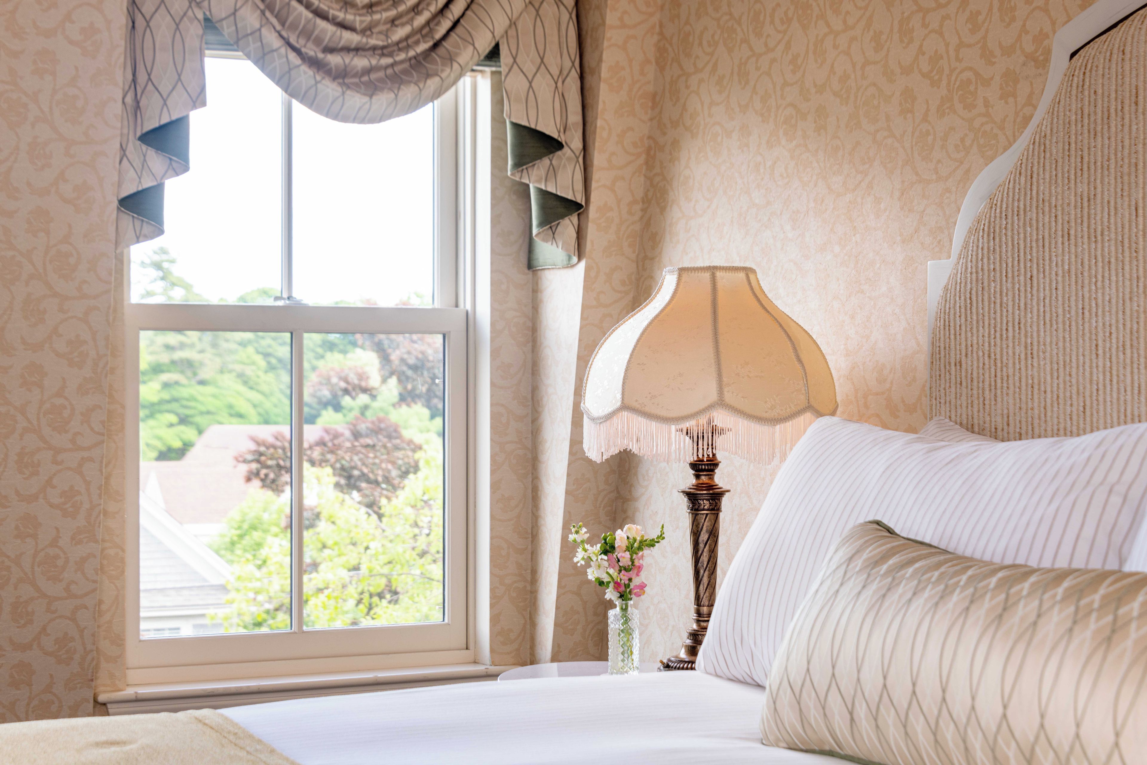 Een hotelkamer met een groot raam, een bed met witte lakens, een decoratieve lamp en een vaas met bloemen.