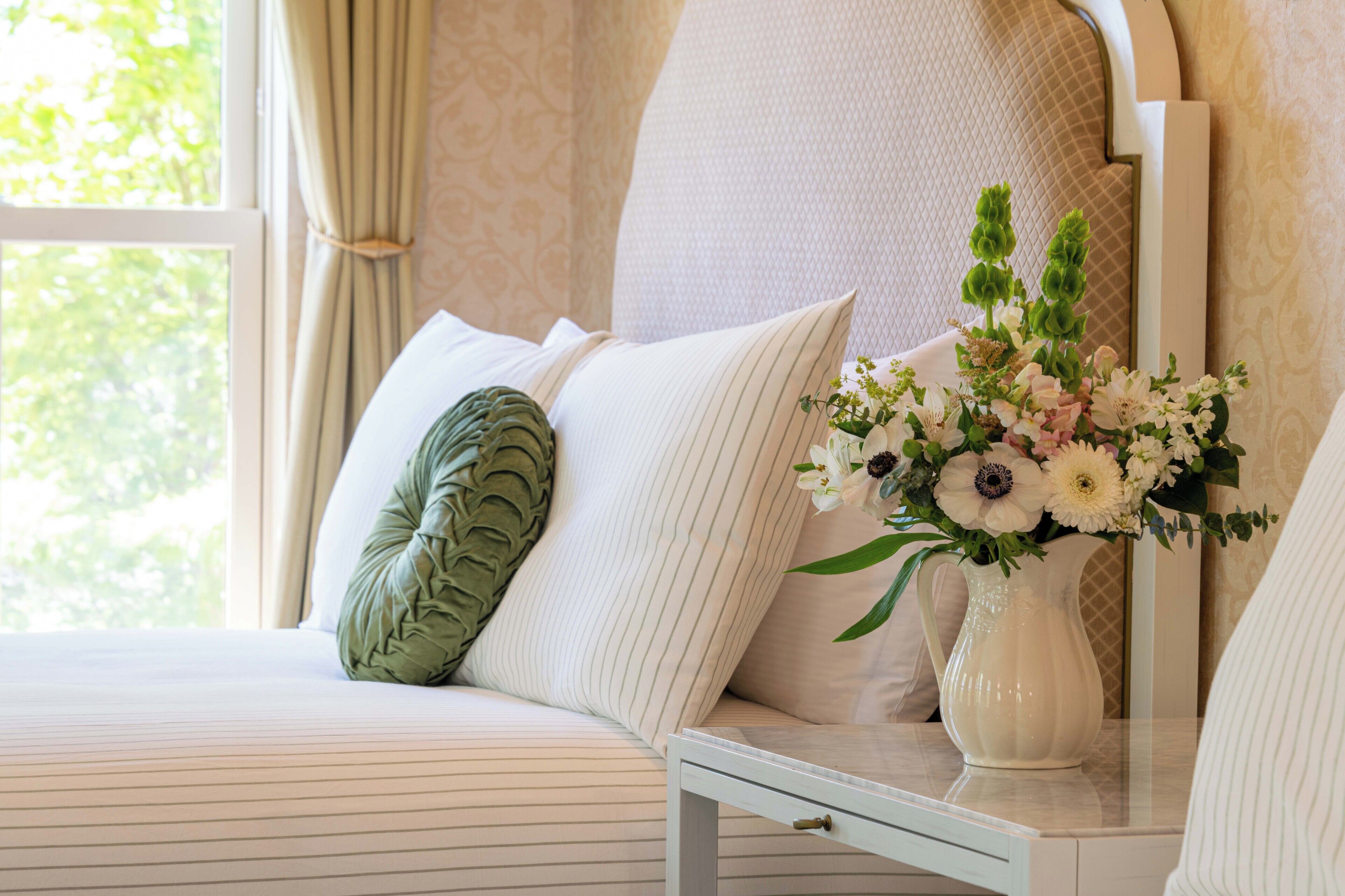 Una cama bien hecha con ropa de cama a rayas, una almohada decorativa verde y un jarrón con flores sobre una mesita de noche.
