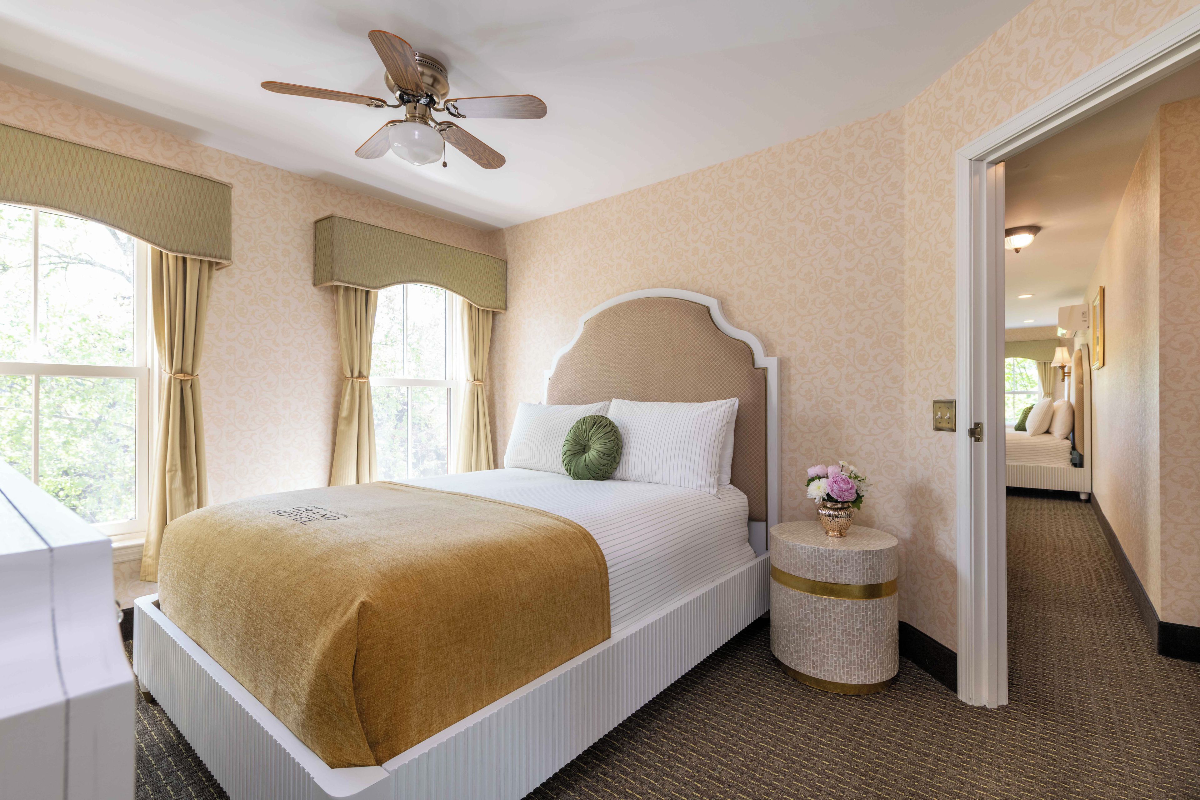 A hotel room with a bed, a round side table, a ceiling fan, and a view of the outdoors through a window.