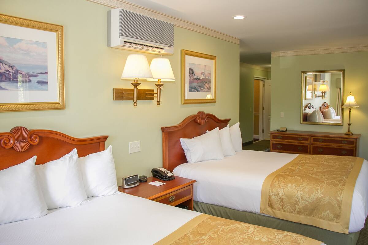 A hotel room with two beds, a wooden headboard, a bedside table with a lamp, a telephone, and a wall-mounted air conditioner.