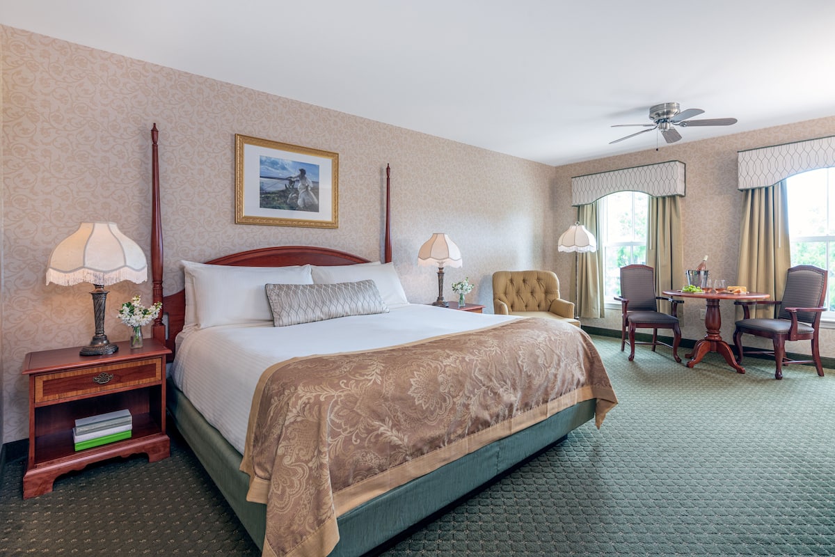 A hotel room with a large bed, two bedside tables with lamps, a chair, a small table with a vase, and a ceiling fan.