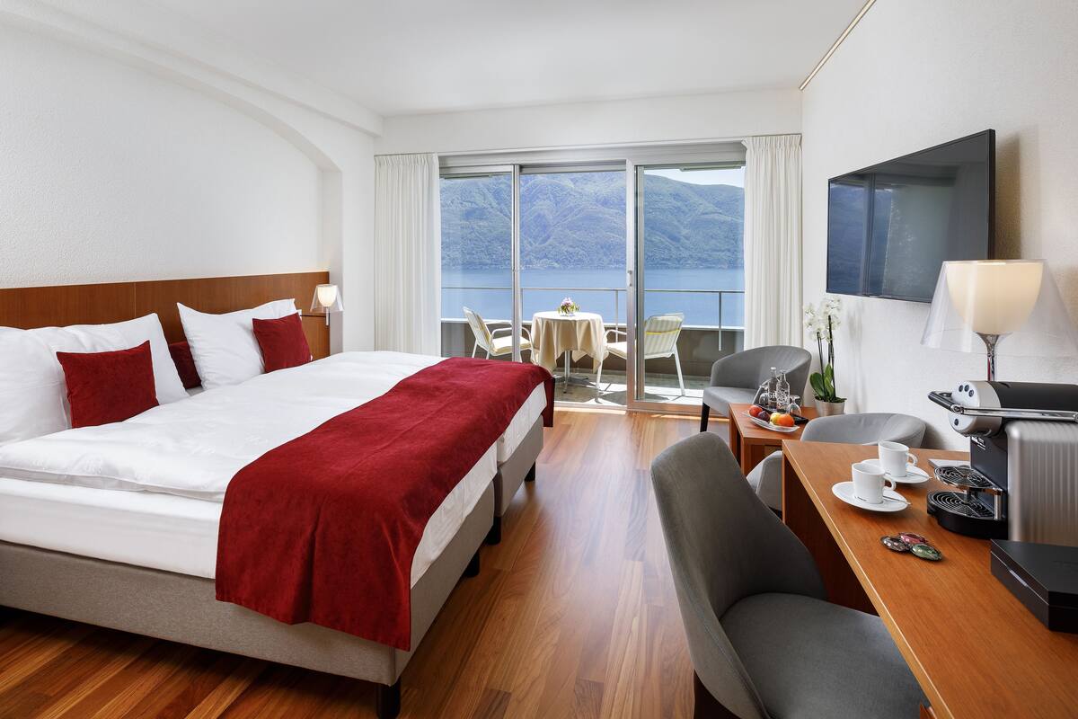 A hotel room with a bed, a desk with a coffee maker, a chair, and a table with cups and a fruit basket.