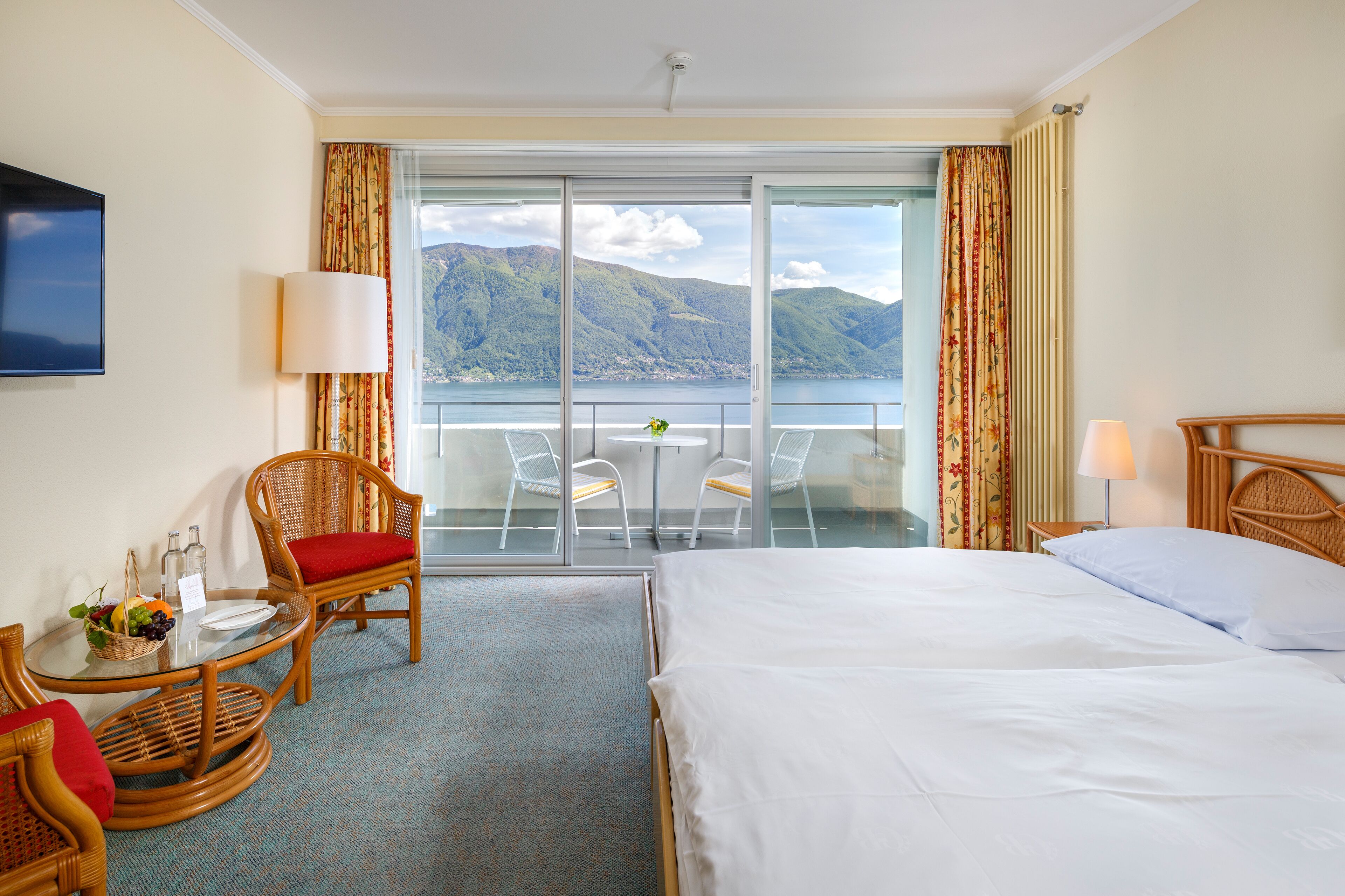 Habitación de hotel con cama, una mesa de comedor con sillas, una mesita con una cesta de fruta y vistas a las montañas y al agua a través de amplios ventanales.