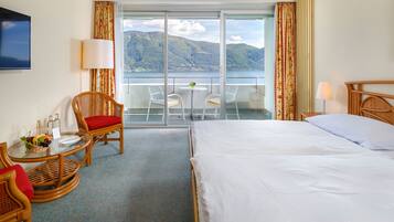 Habitación de hotel con cama, una mesa de comedor con sillas, una mesita con una cesta de fruta y vistas a las montañas y al agua a través de amplios ventanales.