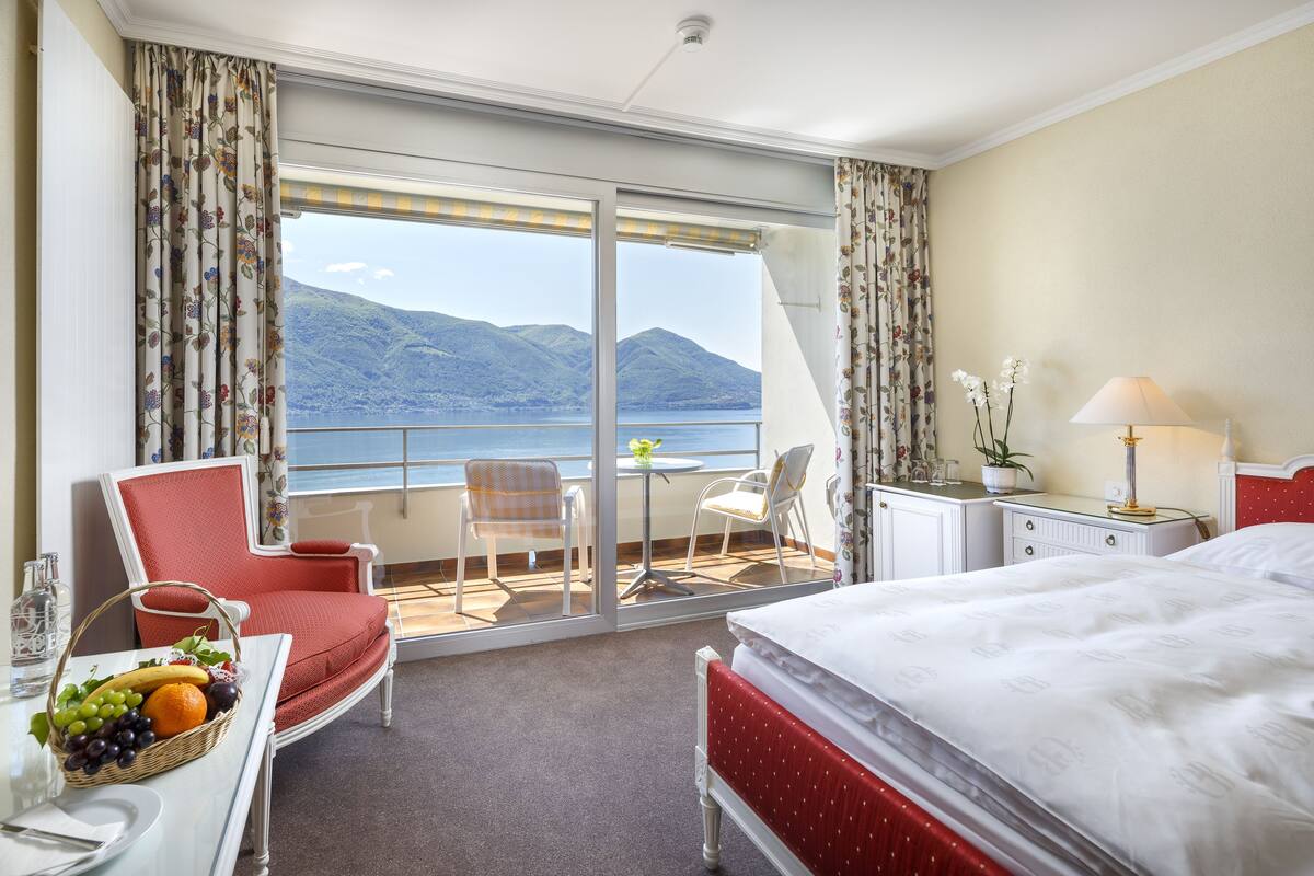A hotel room with a bed, a dining table with fruit, a chair, a small table with a plant, and a view of mountains and water.