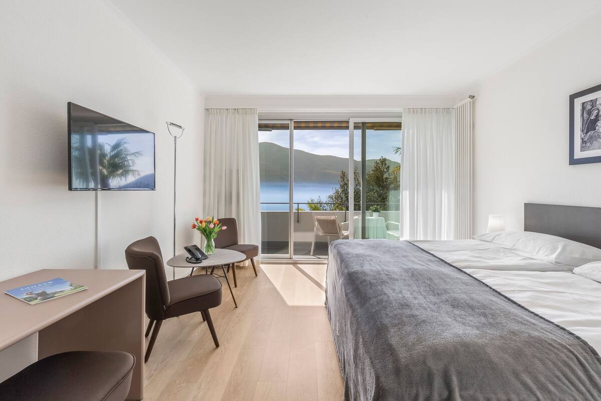 A hotel room with a bed, a desk, a chair, a small table with a vase of flowers, and a view of the sea through a sliding glass door.