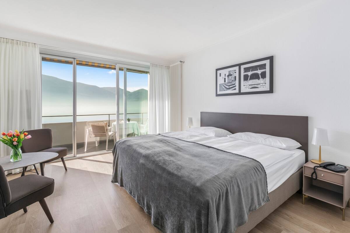 A hotel room with a bed, a nightstand, a desk with a chair, and a view of the mountains through a sliding door.