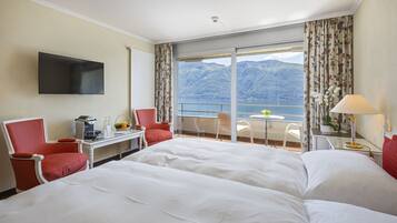 A hotel room with a large bed, a desk with a chair, a flat-screen TV, and a view of mountains and a lake.
