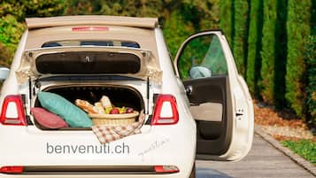 A white car with its trunk open, revealing a wicker basket with bread, apples, and a green cloth.
