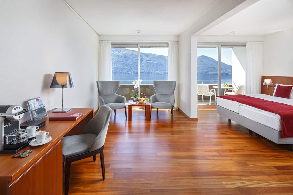 A hotel room with a wooden floor, a desk with a coffee machine, two armchairs, a bed with a red blanket, and a balcony with a mountain view.