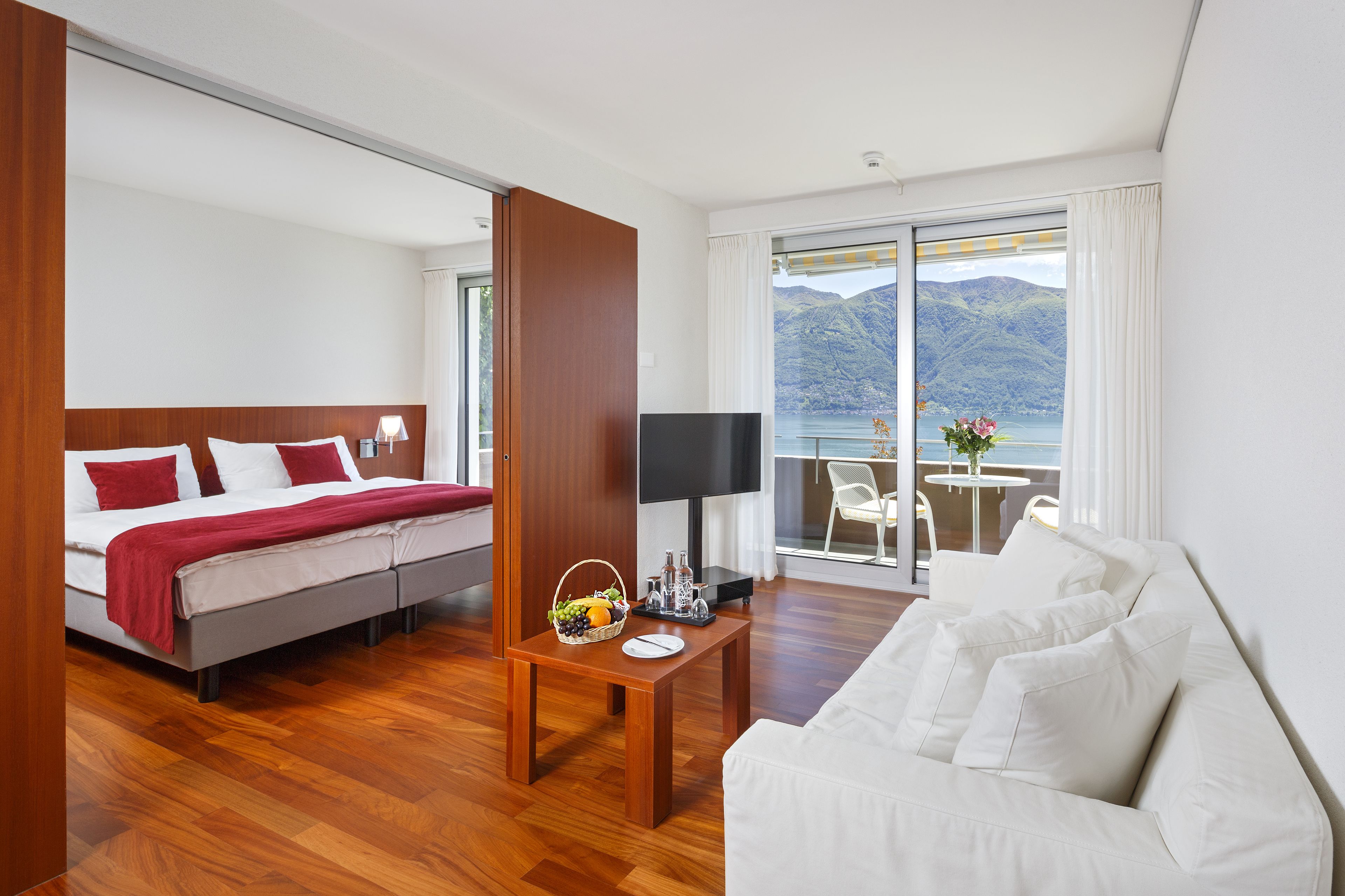 A hotel room with a bed, a sofa, a small table with a fruit basket, and a view of mountains through large windows.