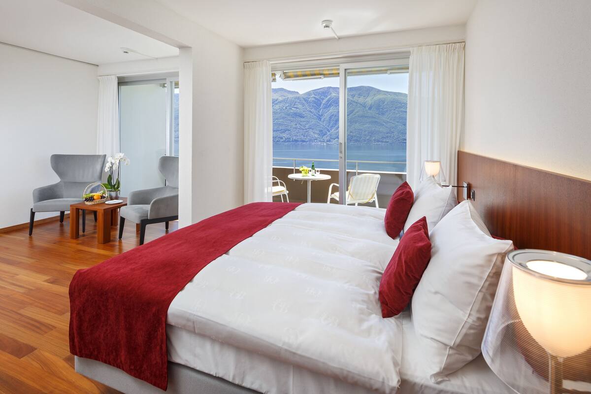 A hotel room with a large bed, a balcony with a view of mountains, a sitting area with chairs, and a small table with a vase of flowers.
