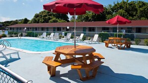 A pool area with wooden picnic tables and red umbrellas, surrounded by white lounge chairs. - Economy Motel Inn & Suites (Somers Point)
