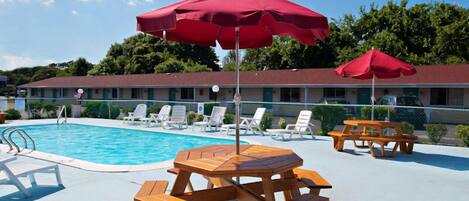 A pool area with wooden picnic tables and red umbrellas, surrounded by white lounge chairs.