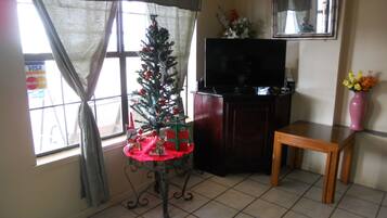 A living room with a Christmas tree, a flat-screen TV, a small table, and a window with curtains.