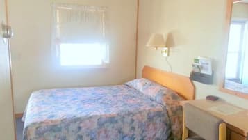 A single bed with a floral bedspread, a bedside table with a lamp, a small desk, and a window with curtains.