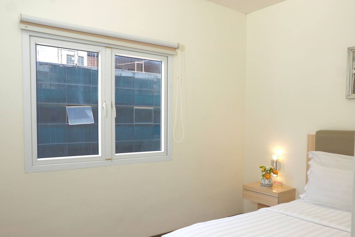 A hotel room with a bed, a nightstand with a lamp, a window with blinds, and a wall-mounted light fixture.
