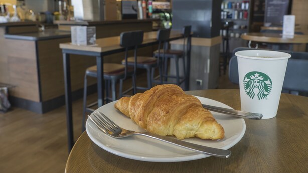 Ein Croissant auf einem Teller mit einer Gabel und einer Starbucks-Tasse auf einem Holztisch in einem Café.