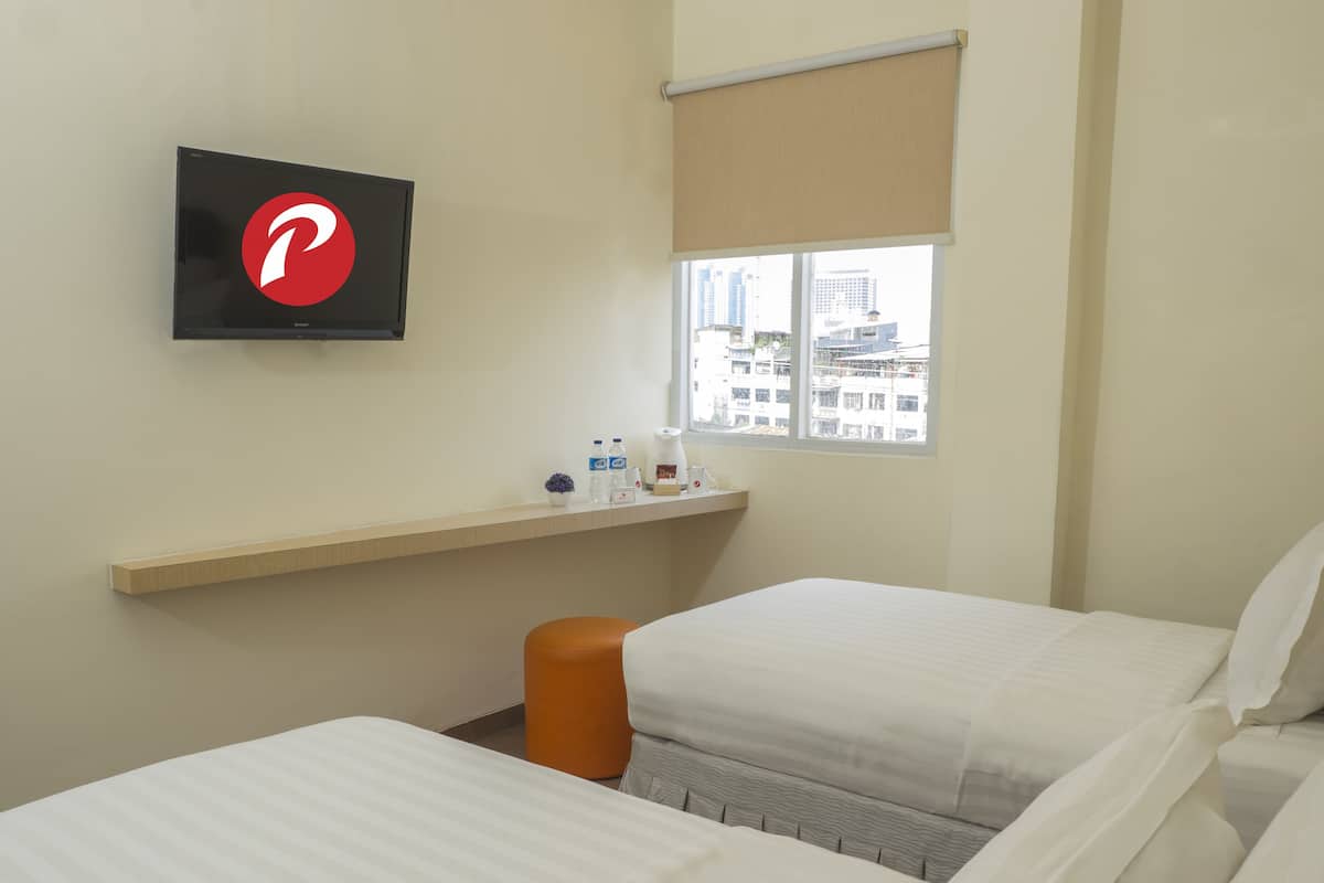 A hotel room with two beds, a TV mounted on the wall, a window with a view of buildings, a shelf with toiletries, and a small orange stool.