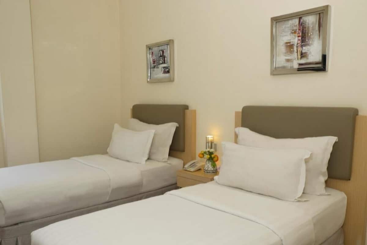 Two single beds with white linens, a bedside table with a lamp, and a small vase with flowers.