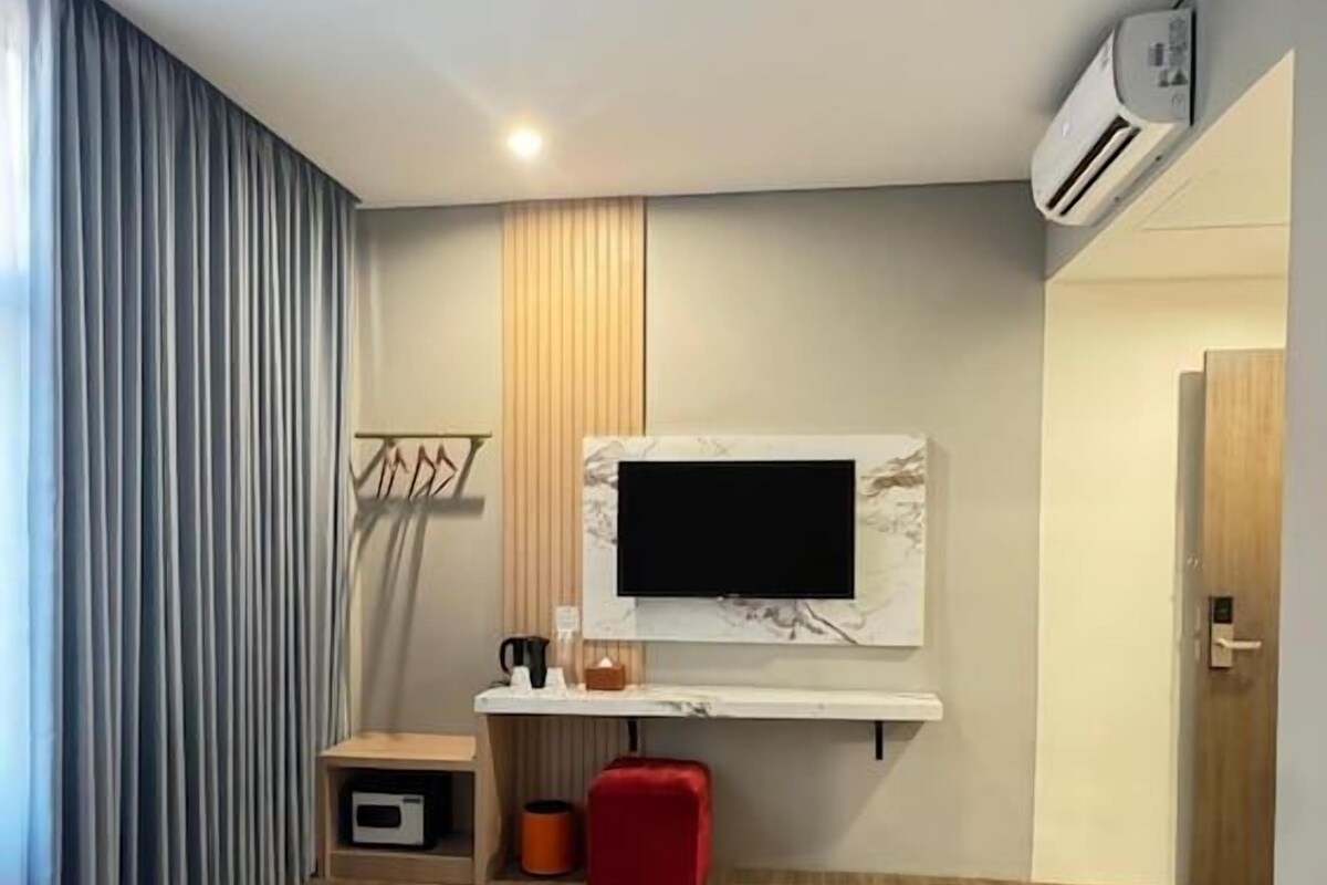 Una habitación de hotel moderna con cama, televisor de pantalla plana en la pared, una pequeña mesa con un taburete rojo y una ventana con cortinas.