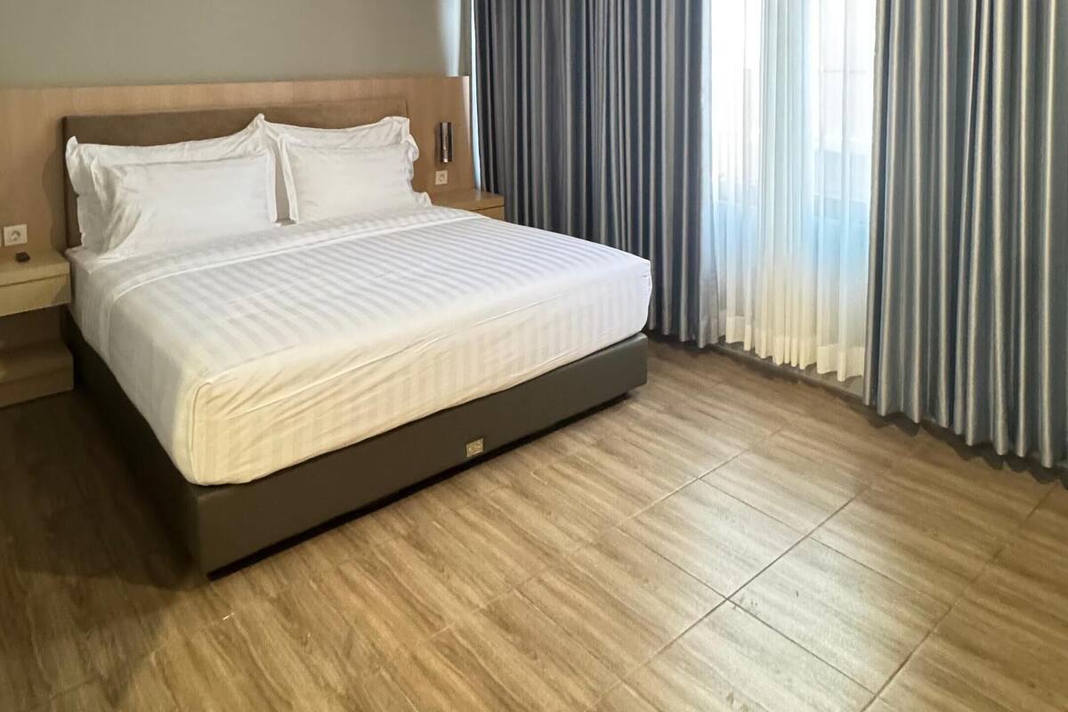 A modern hotel room with a large bed, wooden flooring, and a view of the city through sheer curtains.
